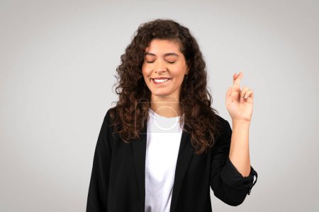 Photo for Hopeful young professional woman in black suit, eyes closed with pleased smile, crossing her fingers for good luck on simple grey background - Royalty Free Image
