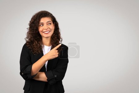 Téléchargez les photos : Radieuse jeune femme d'affaires en blazer noir pointant vers sa droite à l'espace libre avec une expression joyeuse, debout sur fond gris doux, bannière d'affaires - en image libre de droit