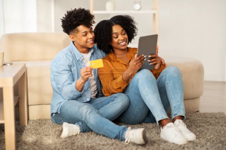 Téléchargez les photos : Niché sur le sol contre un canapé, un jeune couple afro-américain partage une tablette numérique et une carte de crédit, heureux de faire du shopping en ligne ou de planifier dans un environnement chaleureux - en image libre de droit