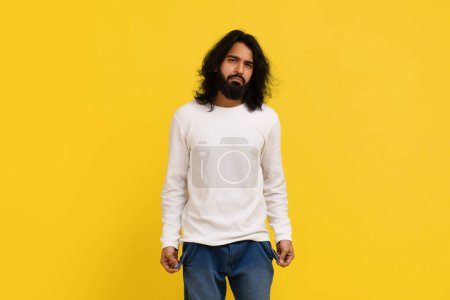 Photo for Poverty and absence of money, permacrisis. Unemployed sad millennial indian man showing empty pockets, posing isolated on yellow studio background, copy space - Royalty Free Image