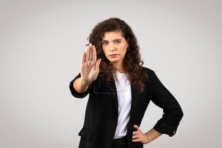 Photo for Assertive young businesswoman in black blazer with stern look, firmly holding up her hand in stop gesture against plain grey backdrop, looking at camera - Royalty Free Image