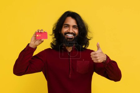 Foto de Feliz hombre hindú milenario peludo mostrando tarjeta de crédito de plástico rojo y pulgar hacia arriba, sonriendo a la cámara, recomendando la banca virtual, el pago sin contacto, fondo de estudio amarillo - Imagen libre de derechos