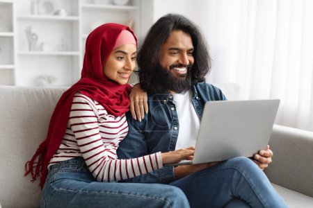 Foto de Sonriente pareja musulmana disfrutando del tiempo junto con el ordenador portátil en casa, cónyuges islámicos felices sentados en un cómodo sofá en el interior de la sala de estar minimalista, utilizando la computadora para el ocio, primer plano - Imagen libre de derechos