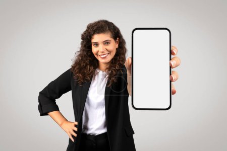 Téléchargez les photos : Femme d'affaires souriante en tenue de cérémonie montrant téléphone portable avec écran blanc, idéal pour les publicités de maquettes d'entreprise, sur fond neutre - en image libre de droit