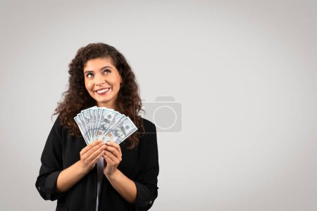 Téléchargez les photos : Femme d'affaires ravie en blazer noir tenant fan de billets de dollar, regardant vers le haut de l'espace libre avec une expression pleine d'espoir sur fond gris minimaliste - en image libre de droit