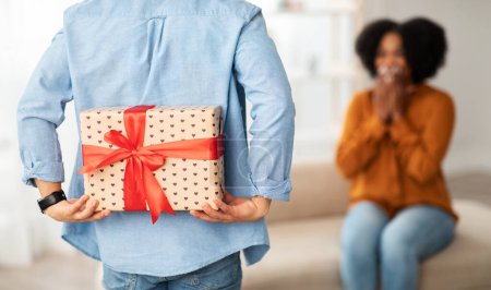 Photo for A african american person from behind holds a large, heart-patterned gift with a red ribbon while a woman in the foreground reacts with excitement and anticipation, cropped, close up - Royalty Free Image