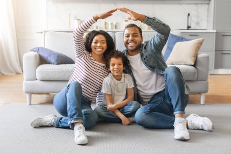 Photo for Family Care. Young black parents making symbolic roof of hands above little son while sitting together on floor in living room, african american mom and dad having fun with their child at home - Royalty Free Image