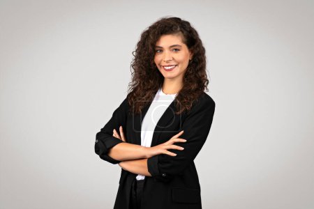 Foto de Joven empresaria europea profesional en blazer negro con sonrisa brillante y brazos cruzados con confianza se levanta sobre fondo gris - Imagen libre de derechos
