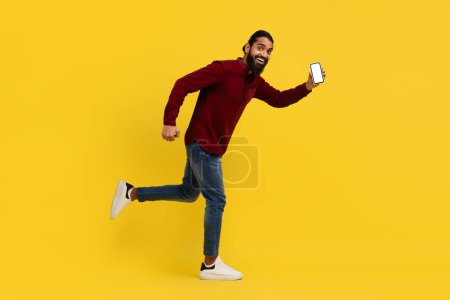 Foto de Date prisa, gran oferta en línea, promoción. Emocionado barbudo hombre indio milenario hipster corriendo con teléfono inteligente con pantalla blanca en blanco en la mano, aislado en el fondo amarillo, espacio de copia - Imagen libre de derechos