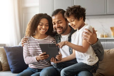 Téléchargez les photos : Portrait de joyeux parents afro-américains et petit fils se détendre avec tablette numérique tout en étant assis ensemble sur le canapé dans le salon, souriant famille noire de trois en utilisant le gadget moderne à la maison - en image libre de droit