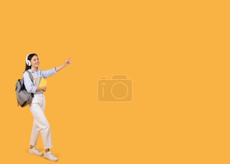 Photo for Smiling young female student with white headphones and backpack pointing to blank space, holding notebooks, on bright yellow background, full length - Royalty Free Image