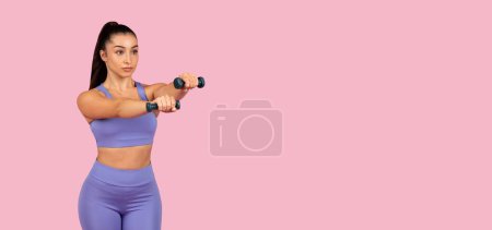 Téléchargez les photos : Jeune femme sérieuse en tenue de sport lavande s'engage dans l'entraînement haltère, démontrant la force du bras sur fond rose minimaliste, panorama, espace libre - en image libre de droit