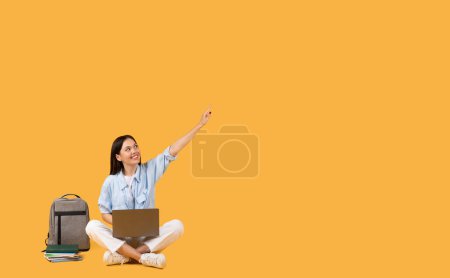Photo for Happy female student in casual wear sitting cross-legged on floor with laptop on her lap, pointing upwards at free space, next to backpack and books on yellow background - Royalty Free Image