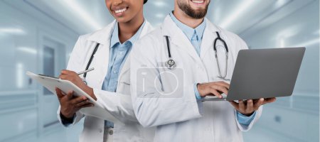 Téléchargez les photos : Un homme et une femme adultes joyeux équipe médicale collaborant dans un hôpital, avec le médecin féminin tenant un presse-papiers et le médecin masculin en utilisant un ordinateur portable à la clinique, panorama - en image libre de droit