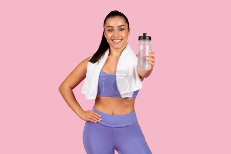 Photo for Fit woman in purple exercise gear smiling and holding water bottle with white towel over her shoulder, signifying post-workout refreshment on pink background - Royalty Free Image