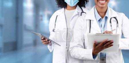 Téléchargez les photos : Deux professionnels de santé dans un environnement hospitalier, l'un tenant une tablette et l'autre avec un presse-papiers, tous deux portant des blouses de laboratoire et l'autre avec un masque chirurgical à la clinique, panorama - en image libre de droit