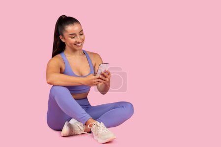 Photo for Relaxed and smiling young woman in trendy activewear takes moment to use her cellphone after rewarding workout session, pink background, free space - Royalty Free Image