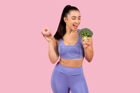 Photo for Smiling young athletic woman in purple activewear deciding between sugary sweet donut and healthy broccoli, standing on pink background - Royalty Free Image