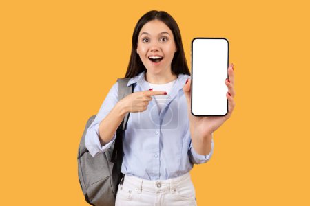 Téléchargez les photos : Jeune femme surprise en chemise bleue, pointant vers l'écran vierge du smartphone avec excitation, portant un sac à dos sur fond jaune, maquette - en image libre de droit
