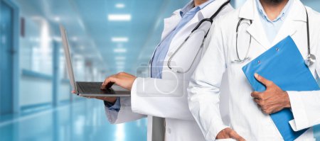 Photo for A pair of medical professionals in a hospital corridor, one operating a laptop and the other holding a clipboard, highlighting the blend of technology and traditional healthcare - Royalty Free Image