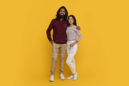 Photo for Cheerful young indian couple posing on yellow background, full length studio shot. Happy loving millennial hindu man and woman wearing casual clothing embracing and smiling at camera, blank space - Royalty Free Image