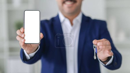 Photo for Close-up of a smartphone with a blank screen held by a smiling mature european businessman in a blue suit, with a blurry background and house keys in his other hand, cropped - Royalty Free Image