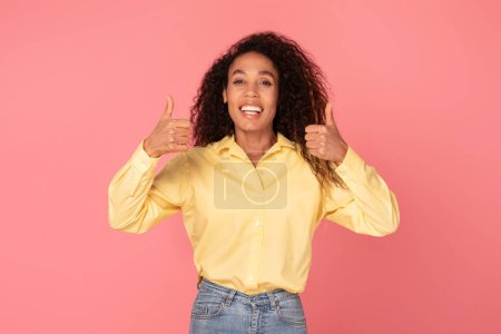 Téléchargez les photos : Femme noire enthousiaste avec les cheveux bouclés donnant deux pouces vers le haut, portant une blouse jaune, exprimant l'approbation et le bonheur sur fond rose - en image libre de droit