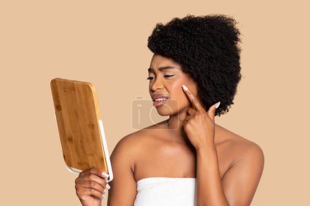 Foto de Joven mujer negra con toalla envuelta, examinando su cara con mirada pensativa usando espejo de bambú, sobre fondo beige neutro - Imagen libre de derechos