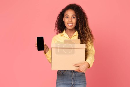 Photo for Stylish young black woman holds box parcels while showcasing phone with blank screen against vibrant pink background, illustrating online shopping, delivery and technology concepts - Royalty Free Image