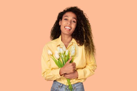 Beaming black woman poses gracefully with bouquet of pristine white tulips on soft peach background, exuding happiness and elegance, conveying joy and beauty