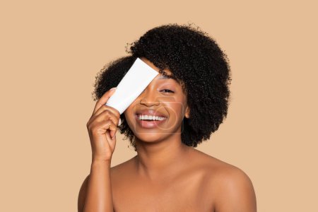 Téléchargez les photos : Jeune femme noire exubérante aux cheveux bouclés couvrant un œil de tube blanc de crème, symbolisant le plaisir et la simplicité des routines de soins de la peau, fond beige - en image libre de droit