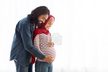 Photo for Loving arab man hugging his pregnant muslim wifes belly while they standing together near window at home, affectionate islamic spouses expecting for a baby, enjoying future parenthood, copy space - Royalty Free Image