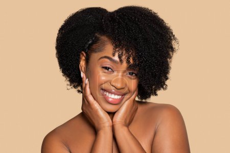 Téléchargez les photos : Délicieuse jeune femme afro-américaine aux beaux cheveux bouclés, les mains reposant doucement sur les joues, donnant un sourire chaud sur fond beige neutre - en image libre de droit