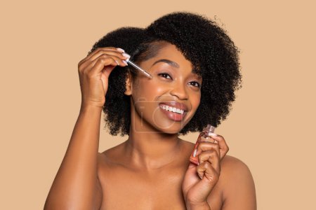 Foto de Joven mujer negra alegre con pelo rizado natural aplicando suero facial para la rutina de cuidado de la piel, sosteniendo la botella del producto, sobre fondo beige suave - Imagen libre de derechos