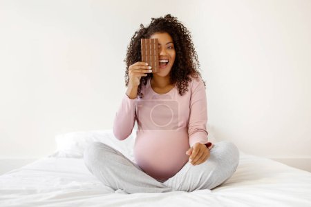 Téléchargez les photos : Femme enceinte noire riante couvrant un œil avec une barre de chocolat tout en étant assise jambes croisées sur le lit à la maison, belle femme enceinte afro-américaine rayonnant de bonheur, espace de copie - en image libre de droit