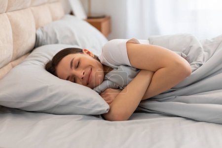 Téléchargez les photos : Jeune femme paisible couchée au lit, embrassant la couverture avec un sourire content, rayonnant de confort et de bonheur dans un cadre serein de chambre à coucher - en image libre de droit