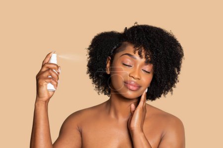 Téléchargez les photos : Femme noire sereine avec les yeux fermés appliquer un spray sur ses cheveux bouclés, vivre un moment d'auto-soin sur fond beige harmonieux - en image libre de droit