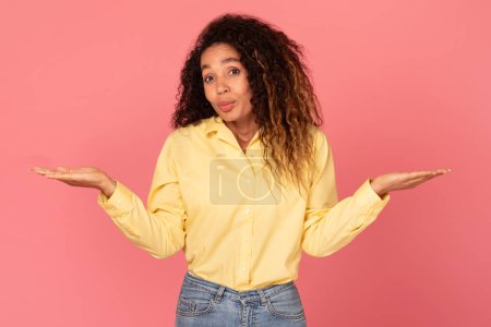 Photo for Perplexed black woman in yellow shirt, hands outstretched in balance gesture, symbolizing weighing options or uncertainty, on pink backdrop - Royalty Free Image