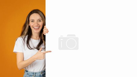 Téléchargez les photos : Une femme millénaire européenne heureuse et rayonnante aux longs sourires de cheveux et pointe vers un espace blanc vide, grande bannière, parfaite pour la publicité, avec un fond orange vif, panorama - en image libre de droit