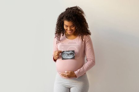 Foto de Concepto de Maternidad. Mujer embarazada negra feliz sosteniendo la foto de ultrasonido de su bebé y abrazando tiernamente el vientre mientras está de pie contra la pared blanca en casa, madre expectante manteniendo la exploración por sonografía - Imagen libre de derechos