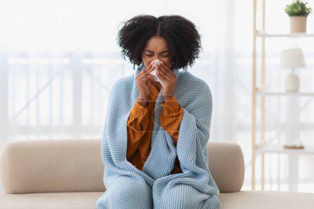 Photo for Young african american unhappy woman feeling sick, wrapped in a cozy blue blanket, sitting on the couch and blowing her nose with a tissue, with a pained expression on her face - Royalty Free Image