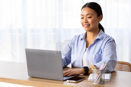 Photo for Chinese woman freelancer working from home office, sitting at table next to window, typing on laptop computer keyboard, sending email, copy space. Remote job, entrepreneurship, online business - Royalty Free Image