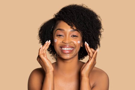 Photo for Cheerful young black woman with curly hair applies white facial cream on her cheeks, smiling with satisfaction at her glowing skin care routine, beige background - Royalty Free Image