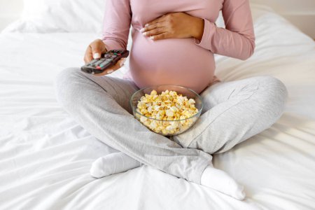 Photo for Cropped Shot Of Pregnant Woman With Popcorn And Remote Controller In Hands Relaxing At Home, Unrecognizable Expectant Mother Watching TV While Resting On Bed In Bedroom And Keeping Hand On Her Belly - Royalty Free Image