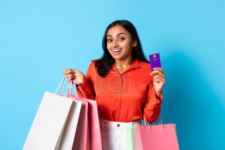 Foto de Compras. Compradora joven india feliz con bolsas de papel en las manos mostrando felizmente su tarjeta de crédito, mostrando emoción sobre la oferta de venta en fondo azul, Studio Shot - Imagen libre de derechos