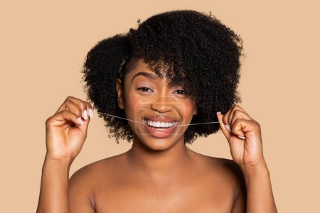 Téléchargez les photos : Femme noire souriante aux cheveux bouclés en soie dentaire joyeuse, promouvant la santé dentaire dans un contexte beige neutre, rayonnant de bonheur - en image libre de droit