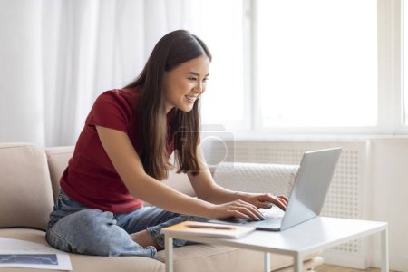 Téléchargez les photos : Focalisé jeune asiatique femme souriant tout en tapant sur ordinateur portable à la maison, heureux coréen femme pigiste confortablement travailler à partir de son lumineux, salon aéré avec décor minimaliste et grande fenêtre - en image libre de droit