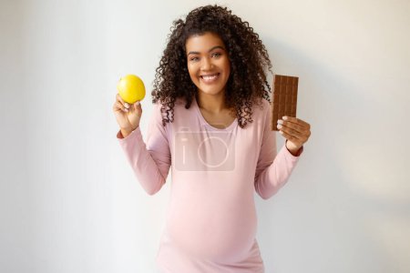 Foto de Mujer embarazada sonriente en rosa superior sosteniendo fruta de manzana saludable y el tratamiento de la barra de chocolate, señora expectante feliz que encarna antojos de embarazo, de pie contra fondo de pared blanca en casa, espacio de copia - Imagen libre de derechos
