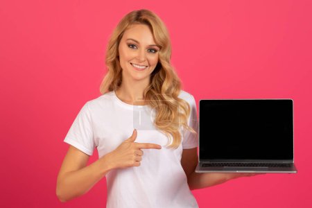 Téléchargez les photos : Heureuse femme blonde pointant vers un ordinateur portable avec écran vide, debout sur fond de studio rose. Lady freelance publicité offre en ligne ou site Web avec ordinateur portable, maquette pour la conception - en image libre de droit