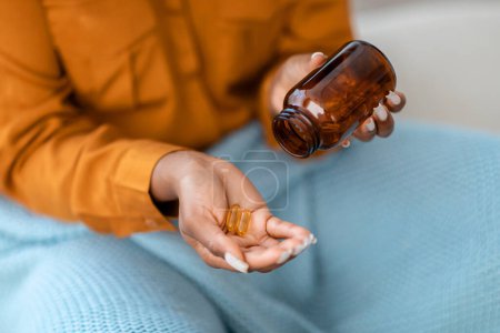 Téléchargez les photos : Une image en gros plan capturant une femme afro-américaine en chemise orange versant des capsules jaunes claires d'une bouteille brune dans sa main, impliquant une prise de médicaments ou un régime vitaminique - en image libre de droit